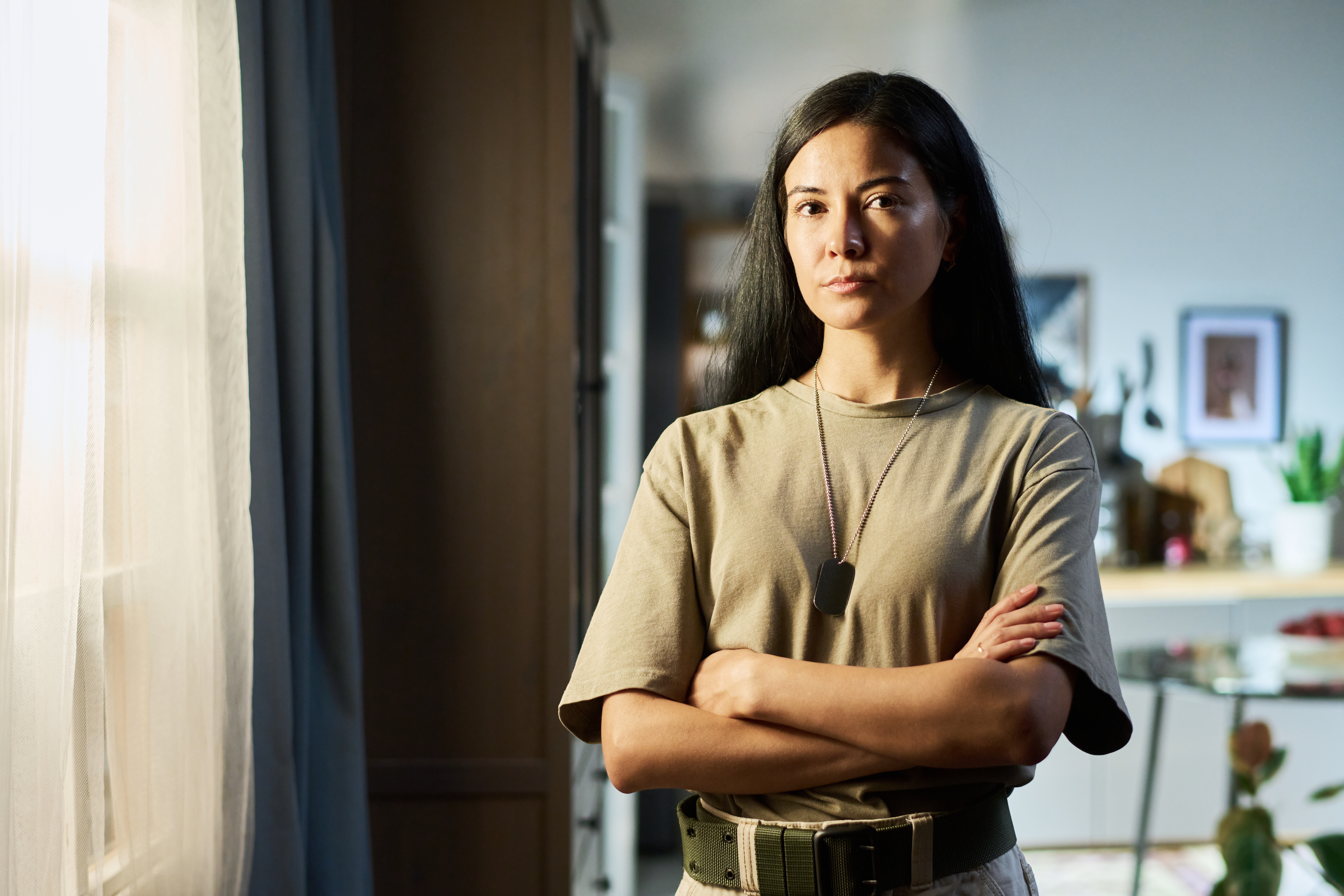 Woman veteran in a tan shirt and dog tags standing at home by a window with her arms crossed